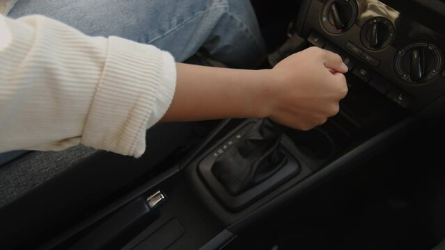 Close-up shot of hand of young dark-skinned female learner in jeans putting gearbox in car with automatic transmission into park position while pressing on brake pedal at end of trip