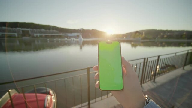 Shot of hands holding vertically phone with green screen moving around. On background lake on sunlight. Nature