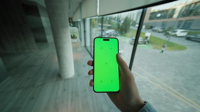Indoors view on hand man holding and scrolling vertically phone with green screen. On background modern space with big windows. Typing on smartphone