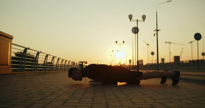 Young athletic man training outside doing push-ups on a city street at sunset. Outdoor workout exercises. Active healthy lifestyle, wellbeing and body care. Slow motion wide shot.