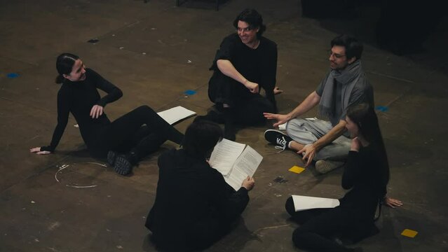 A group of confident young actors in black suits are sitting on stage together with their director holding sheets of paper in their hands with the script of the play on stage in the theater