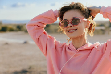 Fashionable woman in sunglasses and pink sweatshirt posing with hands on head in desert landscape