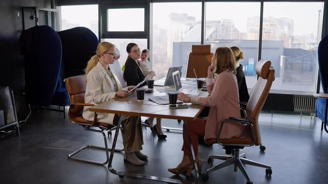 A group of businesswoman with an office desk communicate during a meeting while their colleague a brunette girl in business clothes holds her little infant daughter in her arms in an office with large
