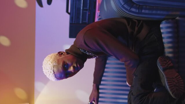 Young creative guy sitting and resting on soft vintage sofa. Trendy cool magnificent african american man looking and posing at camera in retro studio. Disco neon stroboscopes illuminate room.