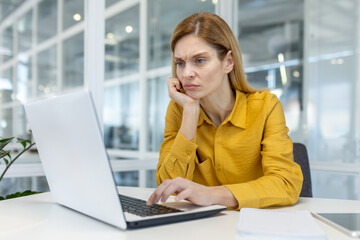 Focused businesswoman in yellow shirt working on a laptop in a modern office environment. Professional and thoughtful expression while concentrating on her work.