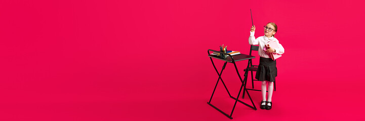Banner. child dressed as teacher, with pointer stick, next to desk filled with school supplies against vibrant pink background. Concept of children dreams, future career, games, fantasy. Copy space.