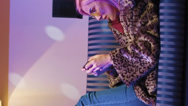 Disco neon stroboscopes illuminate room. Beautiful young creative girl resting on soft vintage sofa. Trendy cool woman in sunglasses laying and playing games online on phone in retro studio.