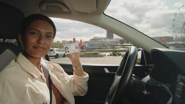 Portrait of happy young African American lady sitting in automobile with seatbelt fastened, holding and showing her drivers license at camera after passing exam