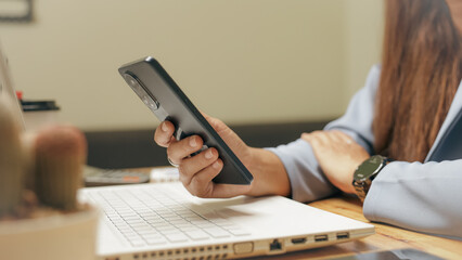 Fototapeta premium A woman is holding a cell phone while sitting at a desk with a laptop. She is focused on her phone, possibly checking messages or browsing the internet. The scene suggests a typical workday