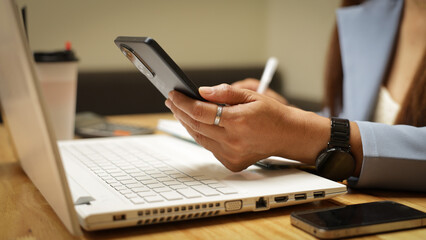 Fototapeta premium A woman is sitting at a desk with a laptop and a cell phone. She is holding a cell phone in her hand and she is typing on the laptop. The scene suggests that she is working or studying