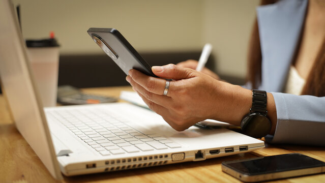 A woman is sitting at a desk with a laptop and a cell phone. She is holding a cell phone in her hand and she is typing on the laptop. The scene suggests that she is working or studying