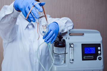 Hands in medical gloves hold out an oxygen mask against the background of the oxygen generator concentrator to deliver at low lung oxygen saturation for Covid-19 pneumonia.