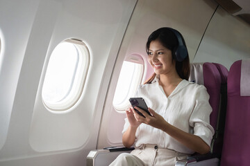 Asian woman using smartphone and headphones on airplane. Concept of air travel, technology, and in-flight entertainment
