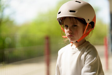 Little cute girl cyclist in a helmet stands next to the fence in the skatepark