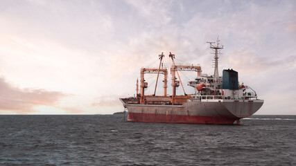 Large bulk carrier ship with cranes sailing on open sea during sunset. The vessel shows signs of wear and tear on its hull. The calm waters and pastel sky create a serene backdrop