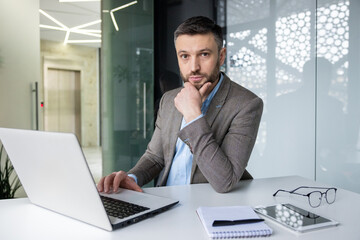 Confident businessman in a modern office setting, using a laptop and contemplating a new business strategy. Professional attire and workspace with natural light.
