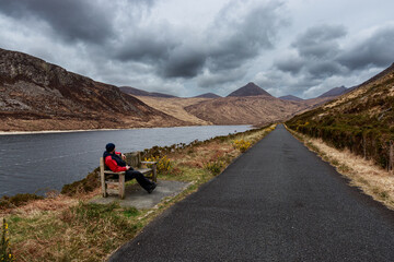 Man in red jacket sit on wood bench lakeshore Silent Valley Reservoir Moure Mountains Lake Northern Ireland UK Europe