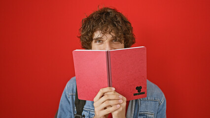 A young man with curly hair peeks over a red notebook against a vibrant red isolated background in a studio portrait.
