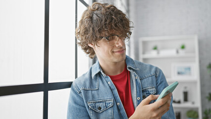 A handsome young man with curly hair wearing glasses and a denim jacket smiles while using a...