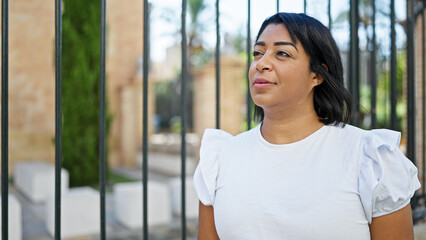 Portrait of a contemplative hispanic woman standing by a garden fence, outdoors in a sunny urban...