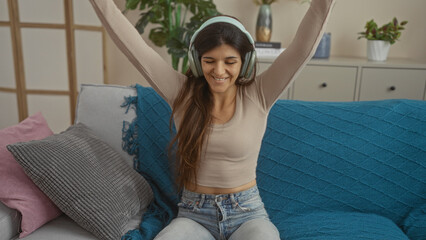 A young hispanic woman enjoying music with headphones, sits in a cozy living room, wearing casual...
