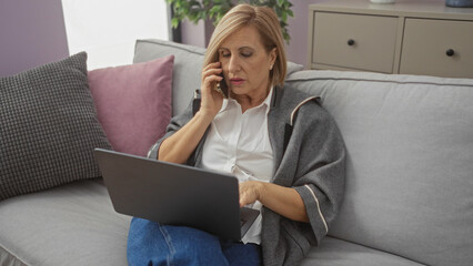 A blonde middle-aged woman sits on a living room couch, speaking on the phone and using a laptop in a comfortable home setting.