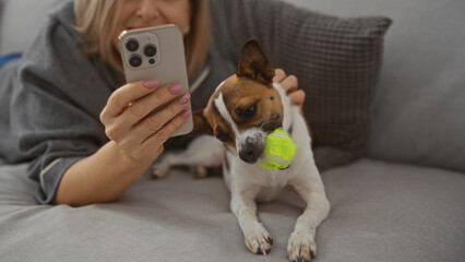 A mature, blonde woman is relaxing in a living room, smiling at her smartphone while her small dog...