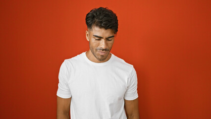 A young hispanic man with a beard stands against an isolated red wall, looking pensive in casual attire.