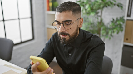 A focused young man with a beard and glasses using a smartphone in a modern office setting.
