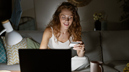 A smiling caucasian woman shopping online with a credit card in a cozy living room with a laptop...