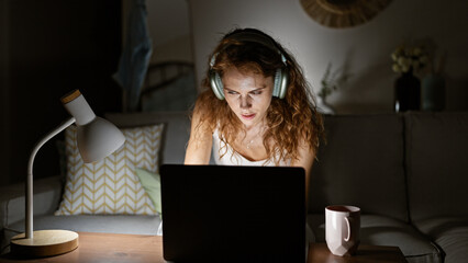 Focused caucasian woman using a laptop at night in a cozy living room with headphones and a desk lamp.