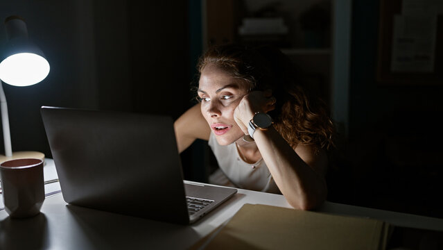Focused young woman working late at her illuminated office desk, staring intently at laptop screen.