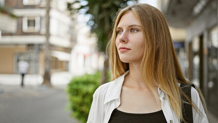 Fototapeta premium Thoughtful young blonde woman with blue eyes, standing on an urban street, wearing casual clothing, looking into distance.