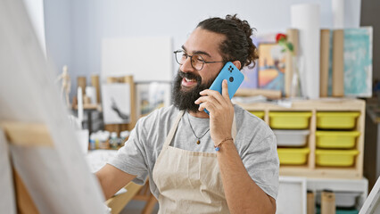 A cheerful hispanic man converses on a smartphone in a bright art studio, embodying creativity and connection.