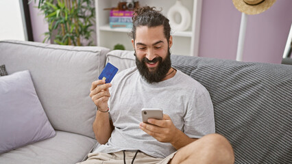 A smiling hispanic man examines a credit card while using a smartphone in a cozy living room setting.