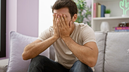Upset hispanic man sitting indoors covering face with hands, showing despair in a living room.
