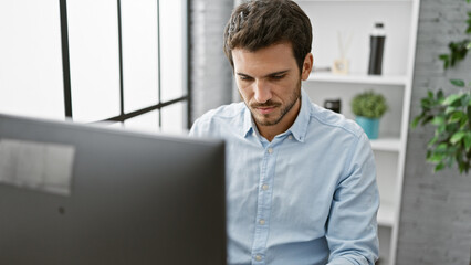 A focused young hispanic man with a beard working on a computer in a modern office interior