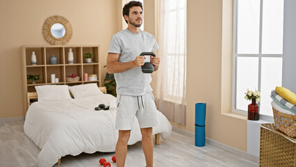 Handsome young man exercising with kettlebell in a modern bedroom setting, implying a personal...