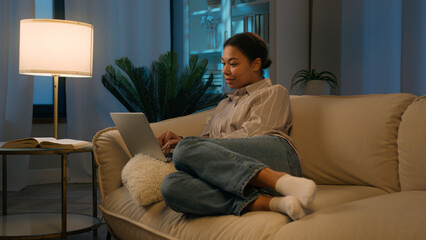 Obraz premium African American woman girl relax on comfortable couch shopping online night time social media addict smiling ethnic female freelancer studying working from home order delivery use laptop application