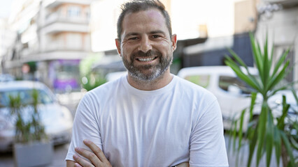 Fototapeta premium Adult hispanic man with grey hair smiles warmly on a sunny urban street, exuding a handsome and approachable vibe.
