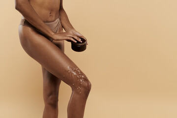 A stylish African American woman with a slim figure poses with coffee scrub on legs on a beige background.