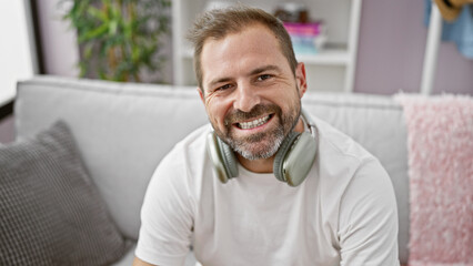 Fototapeta premium Handsome mature hispanic man with grey hair smiling indoors at home on a sofa wearing headphones.