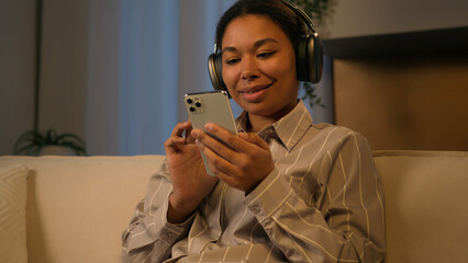 Relaxed smiling African woman in headphones ethnic American girl on home sofa relax comfortable...