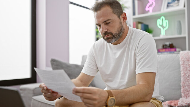 Hispanic mature man with grey hair reading papers in a modern living room, representing everyday home life. - Powered by Adobe