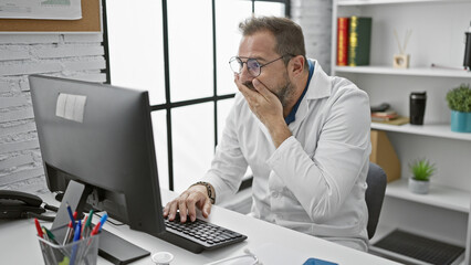 A surprised mature hispanic man with grey hair in a white lab coat works on a computer in a clinic office.