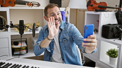 Handsome middle-aged man in jeans jacket waving while making a video call in a home music studio with microphone and keyboard.