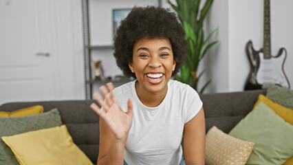 An african american woman with curly hair smiling and waving in a cozy living room setting.