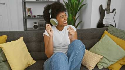 A joyful african american woman listens to music on headphones while relaxing on a gray couch with...