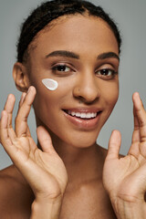 Young African American woman applying cream to her face, focused on skincare routine, exuding...