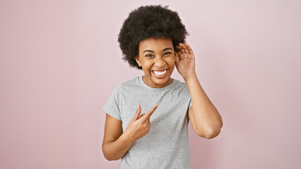 Smiling african american woman with curly hair gestures shush sign against pink background.